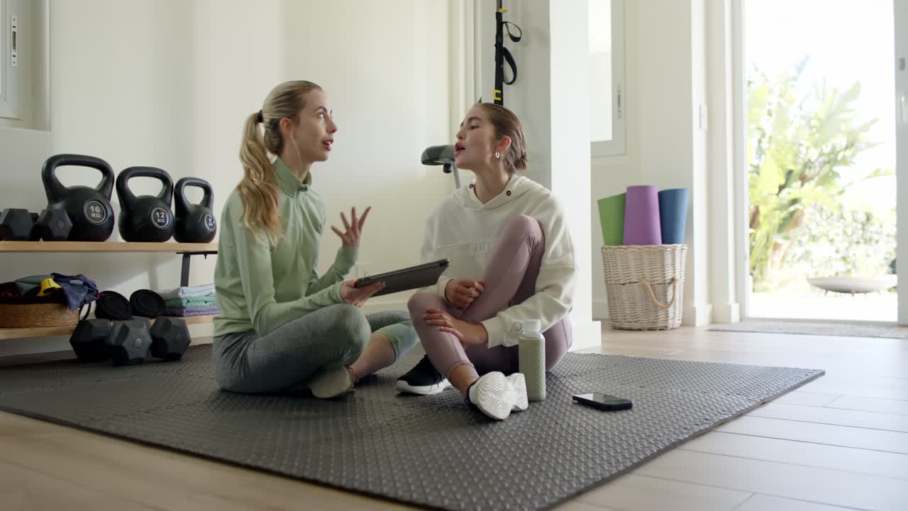 Pelatih wanita mengetuk tablet sementara klien wanita mengangguk untuk memulai tinjauan rencana latihan di atas matras yoga
