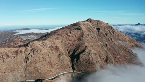 A-rugged-mountain-peak-above-clouds-with-clear-blue-skies,-aerial-view