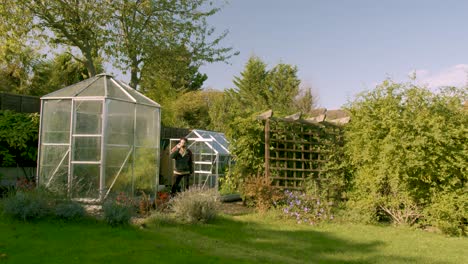 adult male leaves the green house takes a sip from a hot drink and walks through the garden