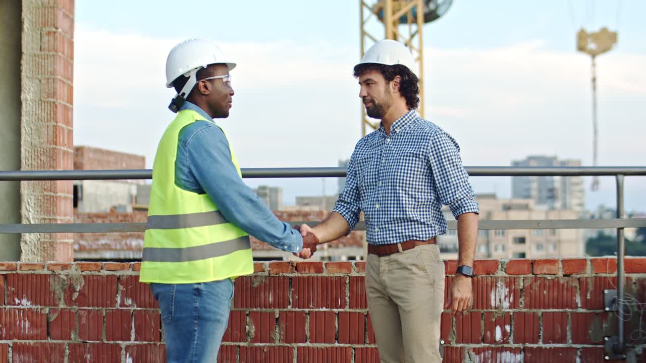 Multiethnic men in hardhats standing at the construcing site, talking and shaking hands. Builder and foreman meeting. Outdoor.