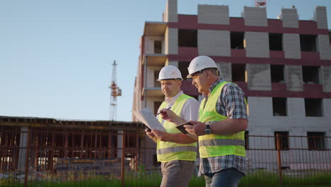 Oberkörperaufnahme-Von-Zwei-Männlichen-Bauarbeitern-Mittleren-Alters-In-Sicherheitskleidung,-Die-Auf-Der-Baustelle-Stehen.-Mann-Benutzt-Walkie-Talkie,-Sein-Kollege-Hält-Papier-Mit-Projektplan-In-Der-Hand