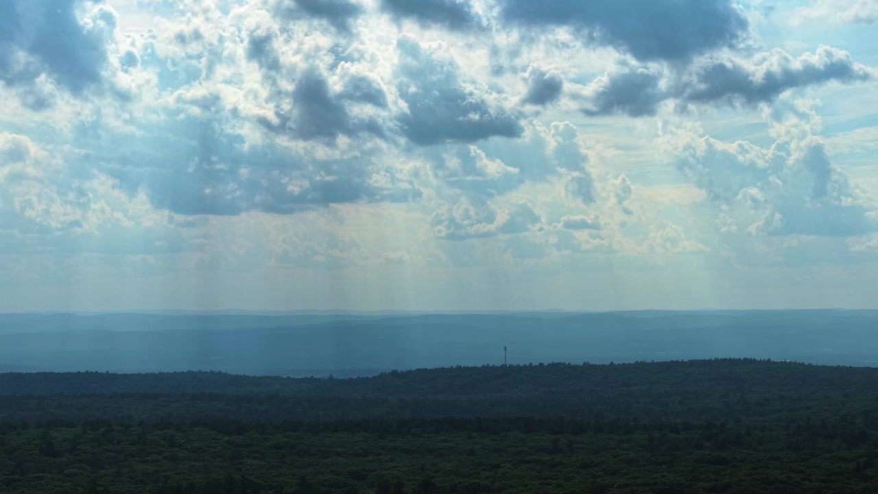 Atemberaubende Weitwinkelaufnahme von Sonnenlicht, das durch strukturierte Wolken strömt und einen üppig grünen Wald darunter beleuchtet. Atmosphärische Beleuchtung schafft eine ruhige, filmische Stimmung, perfekt für Naturthemen.