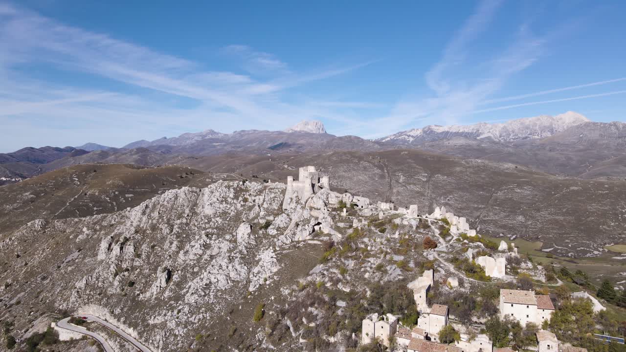 Riprese aeree ravvicinate con drone che orbita attorno alle rovine della fortezza di Rocca Calascio su uno sperone roccioso calcareo, con edifici del villaggio in pietra bianca e il maestoso Gran Sasso innevato sullo sfondo.