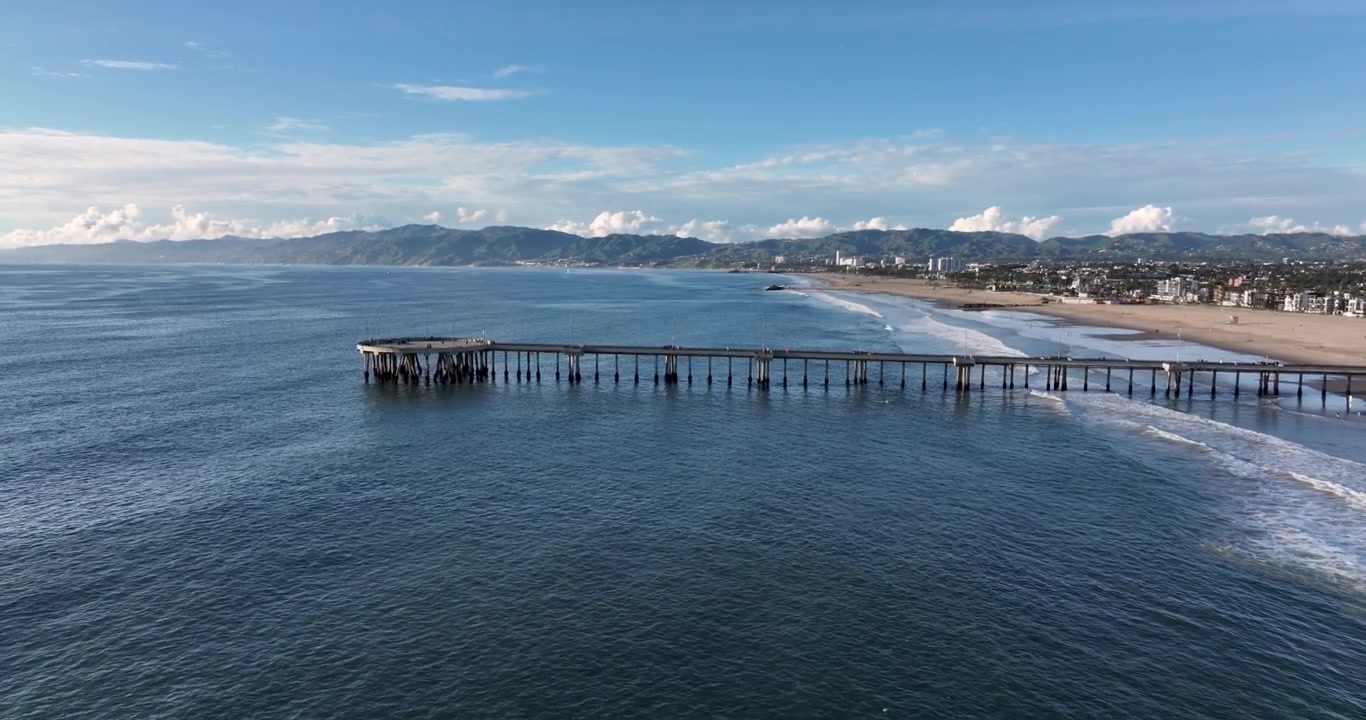 Una toma aérea cinematográfica de gran angular con dron del Muelle de Santa Mónica, sobresaliendo de Venice Beach, girando lentamente en reversa sobre la costa del Pacífico en Los Ángeles, California.