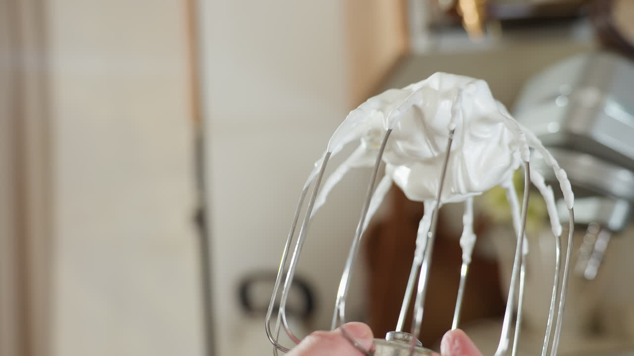 Hand holding metal whisk mixer coated with thick whipped meringue in soft blur kitchen background, highlighting glossy texture and perfect peaks formed during mixing