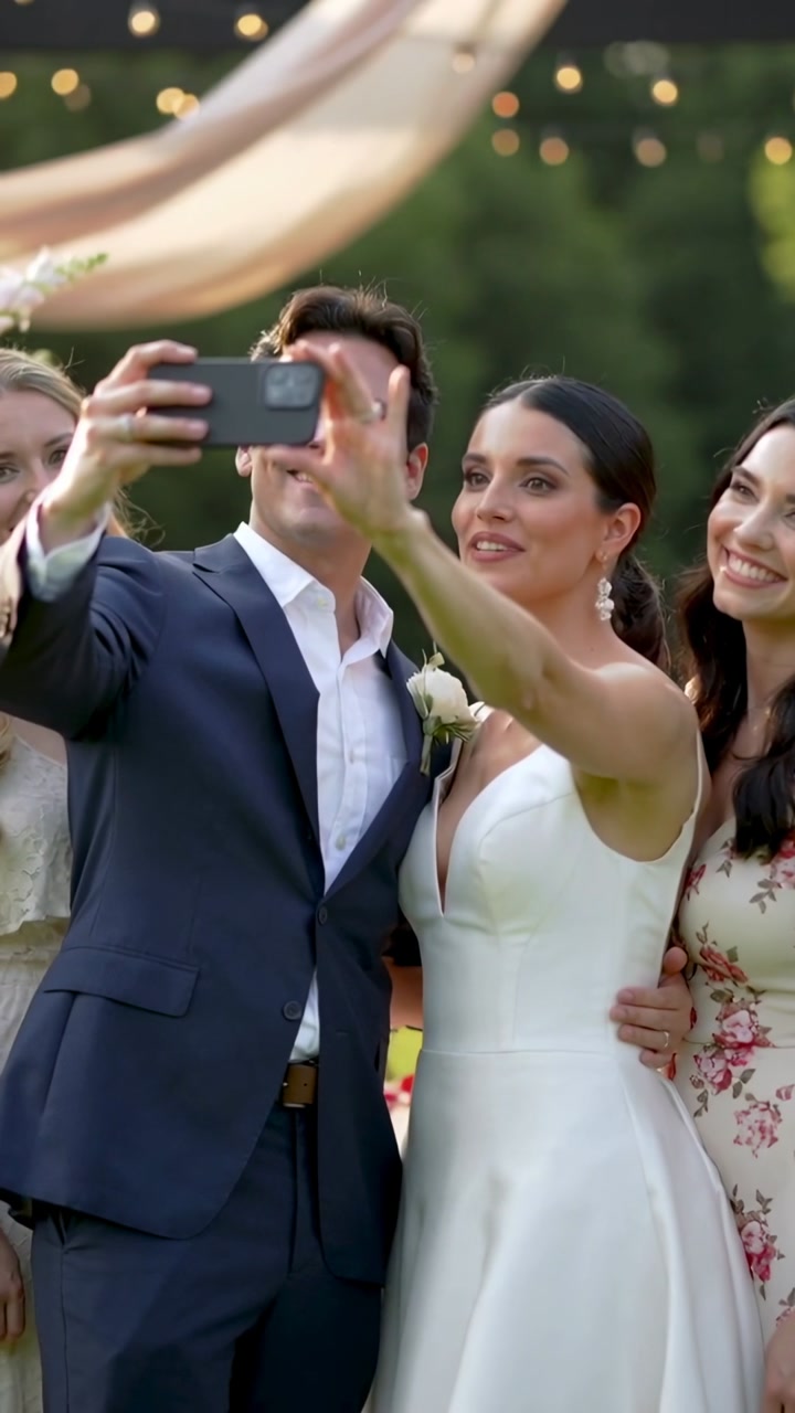 Groom holding phone, bride reaching, friend leaning, smiling, taking wedding selfie under lights