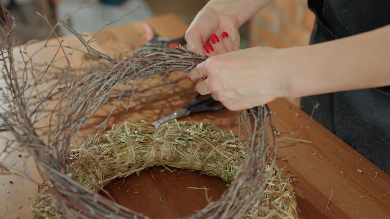Female florist hands carefully shaping rustic wreath by arranging dry twigs on straw base, highlighting handmade craft process on wooden table for seasonal decoration, floral design project indoors