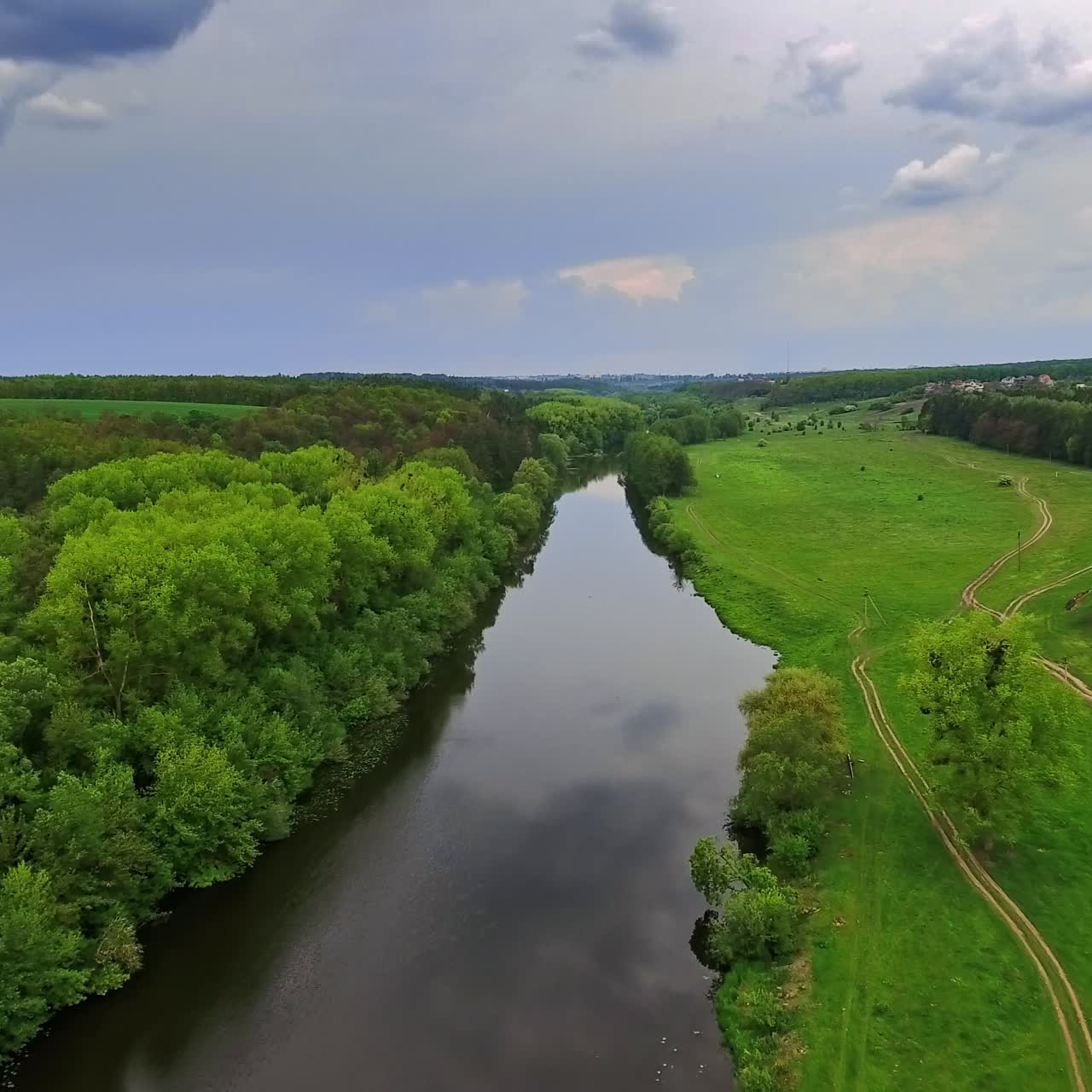 Narrow river with meadow on one side and forest on another. Cloudy sky reflecting in the water. Top view