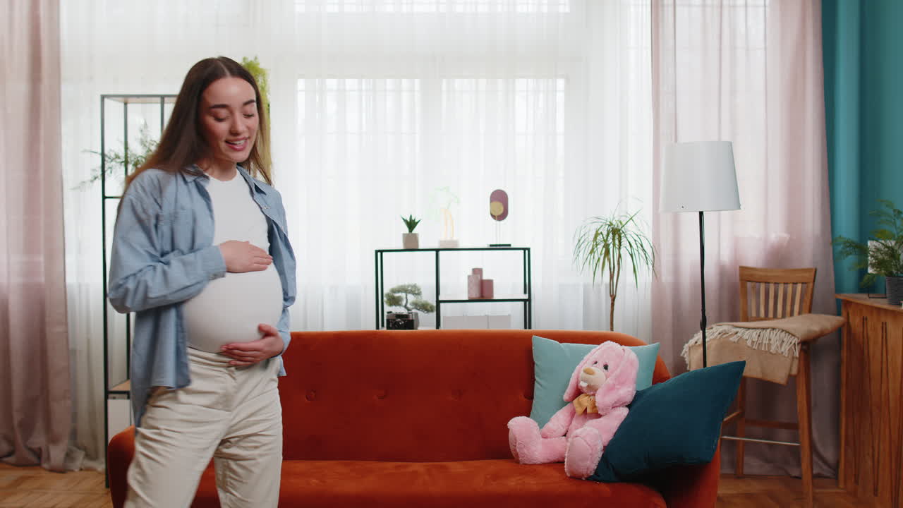 Happy smiling young pregnant woman playfully dancing listening music touching her big belly at home
