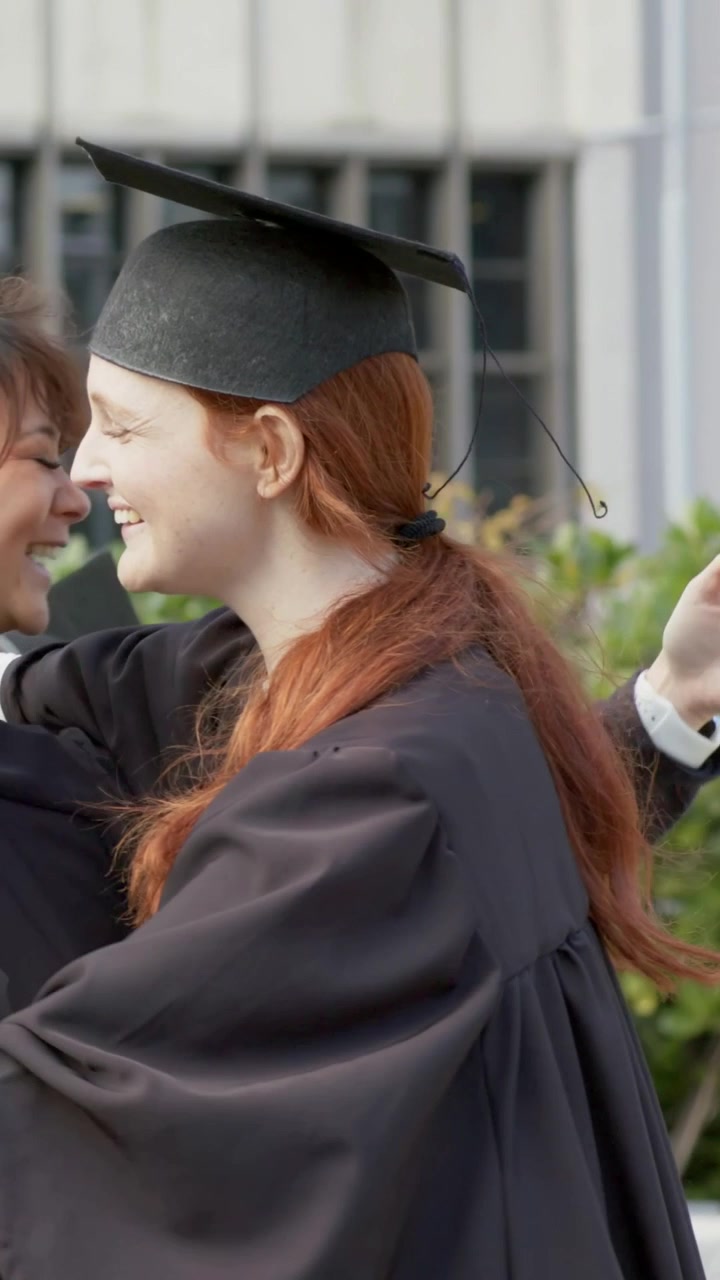 Video verticale: Laureata dai capelli rossi e radiosa che alza il diploma e abbraccia gli amici nel campus, con toge e tocco neri