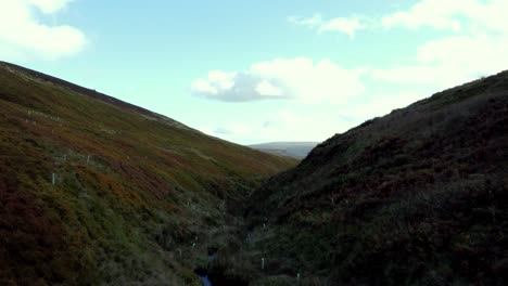 travelling drone shot of a lush green valley with a stream running down the middle with a very old rock formation at the bottom