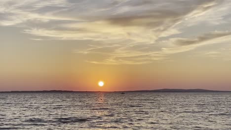 Orange-sun-over-wavy-waters-during-summer-sunset-and-small-boat-passing