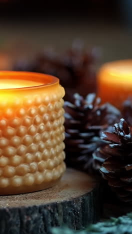 three candles are lit on a wooden table with pine cones in the background. the candles are yellow and the pine cones are brown. scene is warm and cozy, with the candles providing a soft