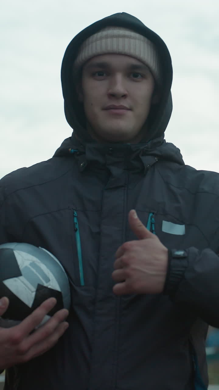 Confident athlete holding soccer ball and giving thumbs up during training in urban arena, showing determination with modern residential buildings in background during cloudy evening