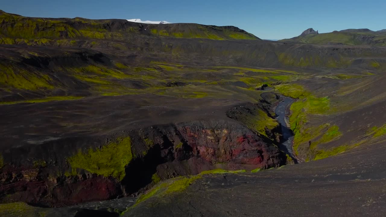 Aerial drone footage flying towards Iceland mountains and rough rocky terrain green and brown landscape while a narrow river is flowing in the middle fast during a sunny day with blue sky in the back.