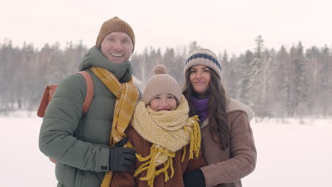 front view of father, mother and daughter in winter clothes hugging and looking at camera in a snowy forest