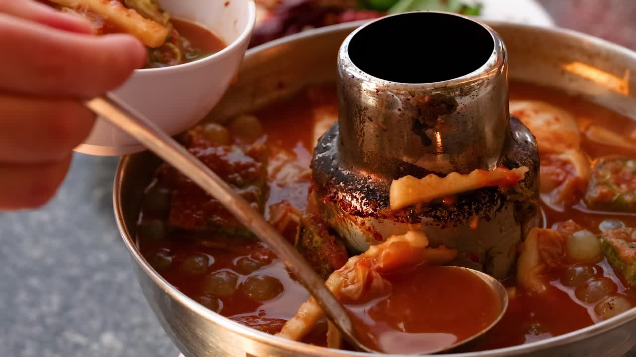 A hand uses a ladle to serve spicy soup from a steaming hot pot into a bowl.