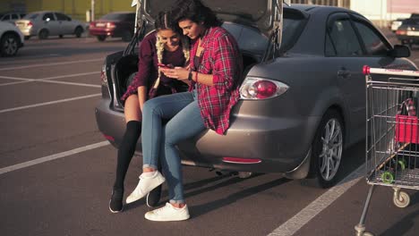 two young attractive girlfriends sitting inside of the open car trunk in the parking by the shopping mall and talking, looking