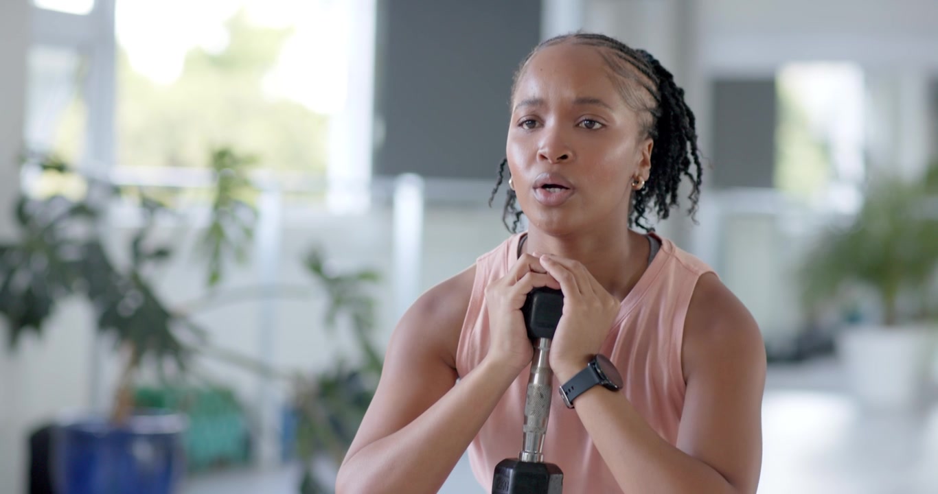 Afrikansk kvinde holder en sekskantet håndvægt og laver goblet squats i et fitnesscenter, iført pink leggings, for at opbygge styrke.