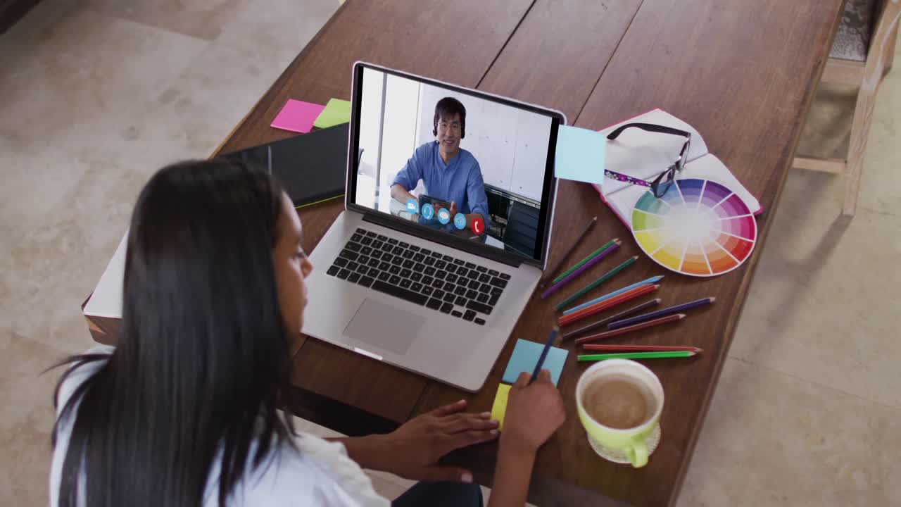 mujer caucásica usando una computadora portátil en una videollamada con un colega masculino y tomando notas trabajando desde casa