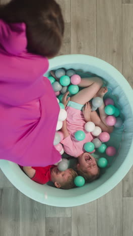 delighted children siblings lie in dry pool at home. caring mother pours out colorful plastic balls from bright bag while boy and girl laugh joyfully