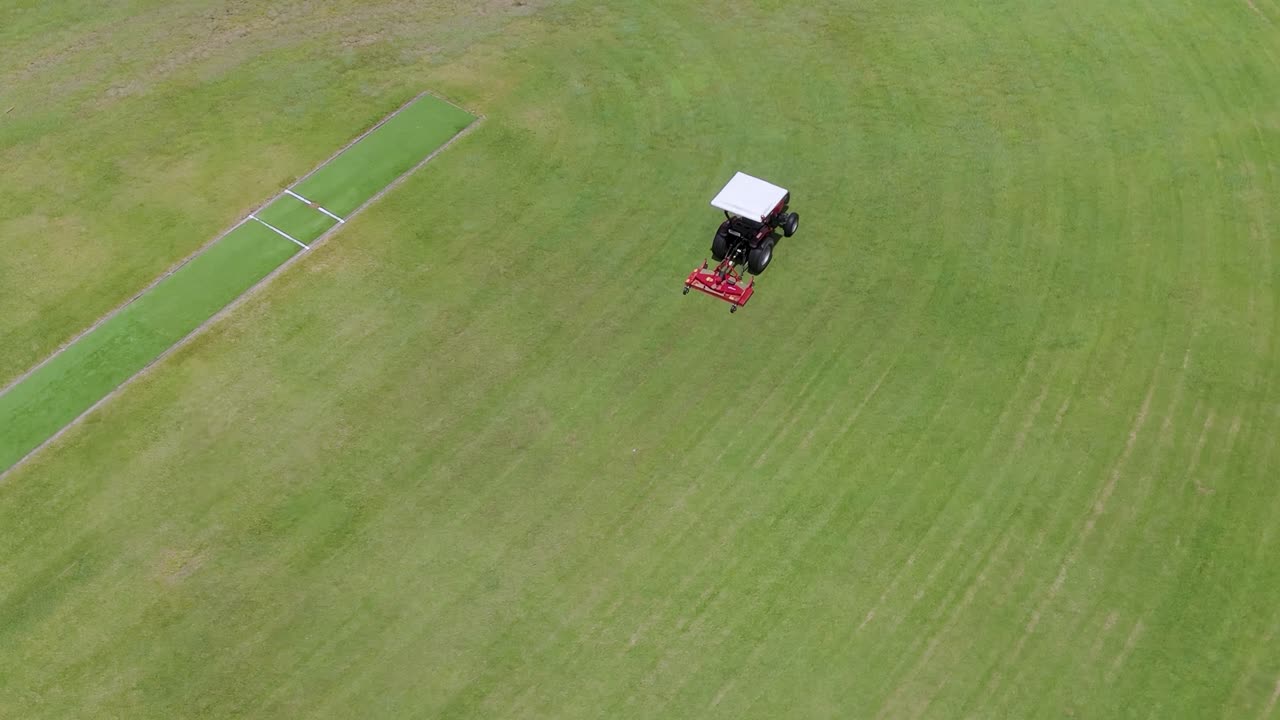 Aerial view of a tractor mowing a green field, creating precise lines and patterns.