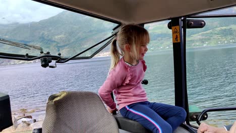 farmers cute young daughter sitting inside tractor close to hardangerfjord sea - eager to learn and start driving tractor - handheld inside tractor cabin - norway