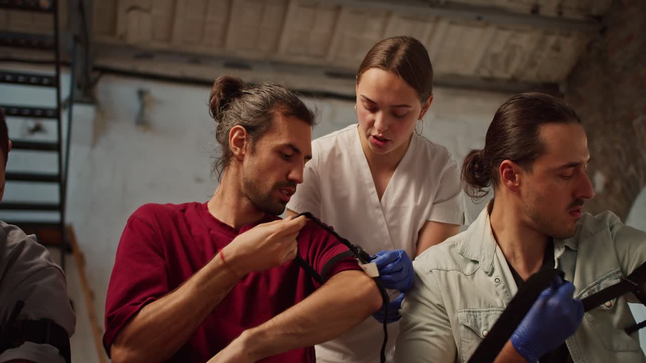 disparo de primer plano: una enfermera profesional con un uniforme médico blanco ayuda a un hombre moreno con cabello largo a poner correctamente un torniquete en su hombro durante un entrenamiento práctico en medicina