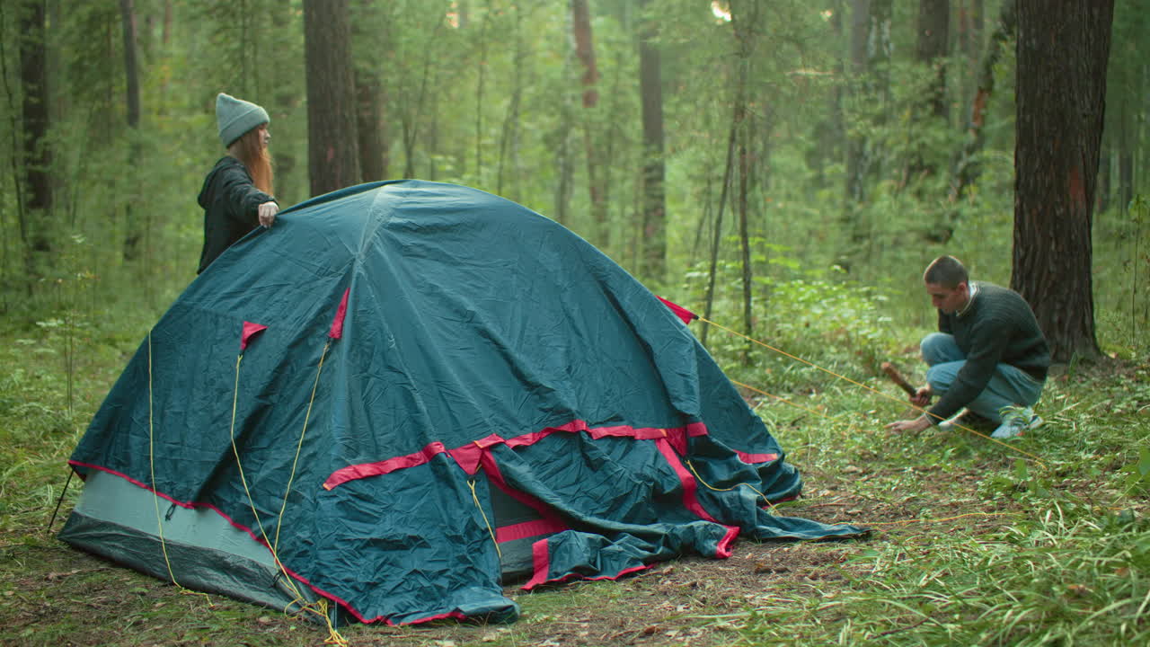 Lady stands beside blue and red tent watching boyfriend crouch and fasten rope to ground with peg in serene forest setting, both involved in calm tent setup