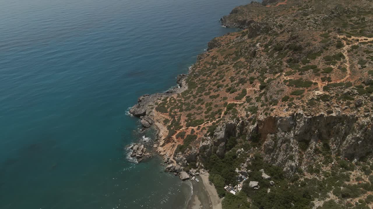 Aerial view of the rocky coastline near Preveli beach in Crete, Greece, showing rugged cliffs descending into a clear blue sea with sparse vegetation on a summer day