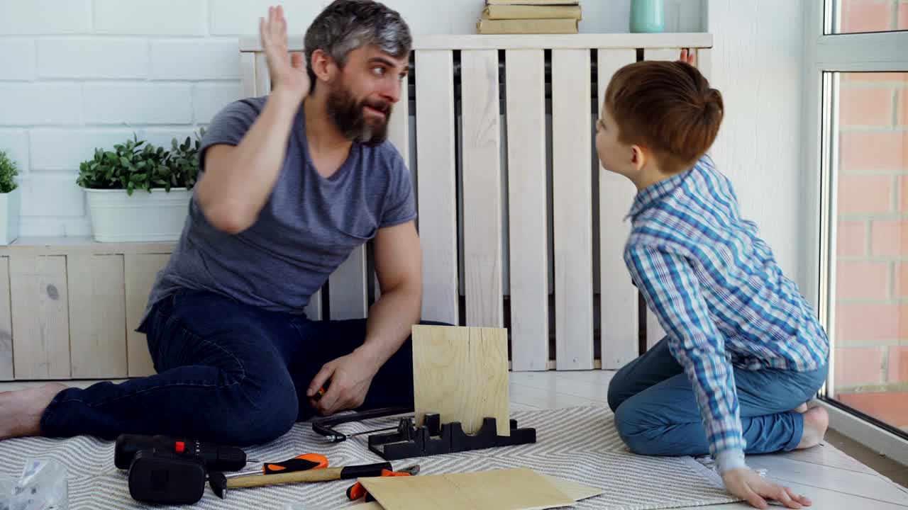 el hombre barbudo está midiendo una hoja de madera con un carrete de medida mientras hace un trabajo de construcción con su hijo y luego hace un high-five para celebrar el trabajo exitoso.