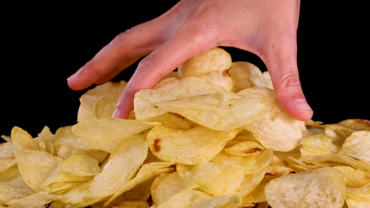 Human hand reaches into large pile of potato chips, isolated against black, with even lighting