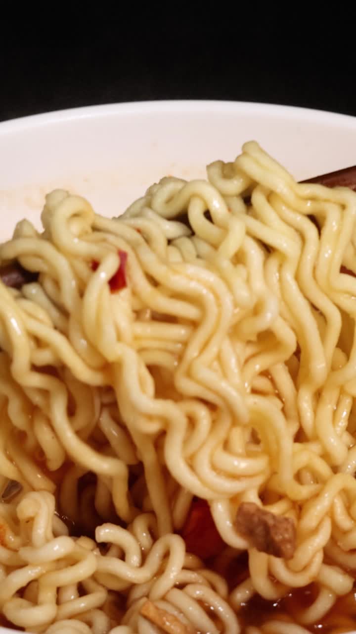 Le bacchette sollevano noodles istantanei da una ciotola di brodo rosso caldo, con illuminazione da studio in primo piano.