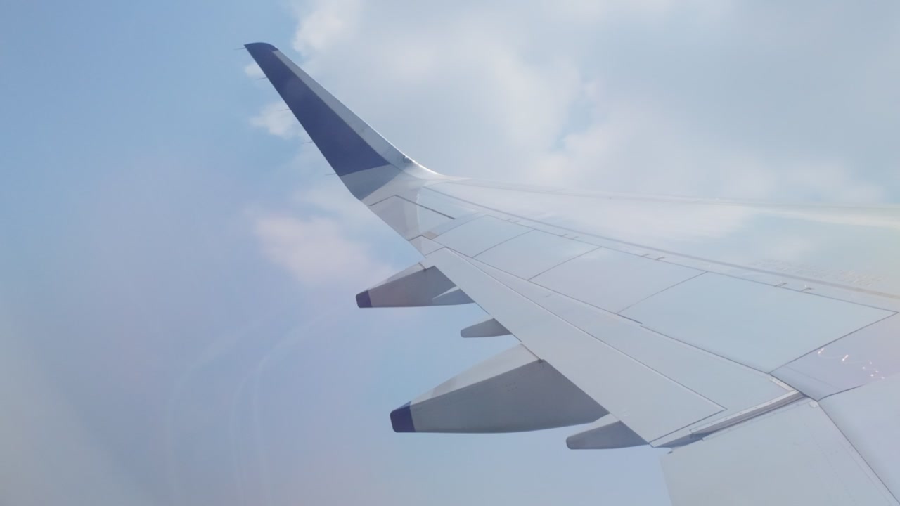 飛行中に雲が流れていく中、激しい乱気流時の飛行機の翼が乗客の窓から見える。澄み切った青空と散らばる白い雲が見える安定した巡航高度。