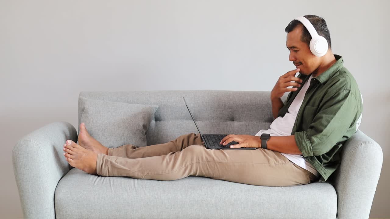 A side view of an Asian man lying comfortably on a grey fabric sofa, wearing white headphones and using a laptop on his lap. He is barefoot, smiling, and has one hand on his chin in a thoughtful pose