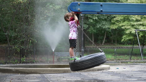 a little boy wearing a face mask stands alone on a tire swing in an empty playground.