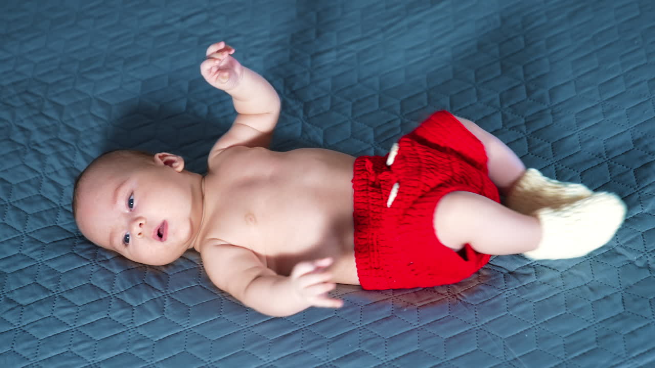 Active toddler boy in red shorts and white socks. Caucasian baby moves feet cheerfully while lying on the bed. Grey backdrop.