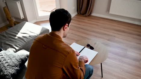 Back-View-Of-A-Blind-Man-Sitting-On-The-Sofa-And-Reading-A-Braille-Book,-Then-Wearing-Glasses-And-Standing-Up