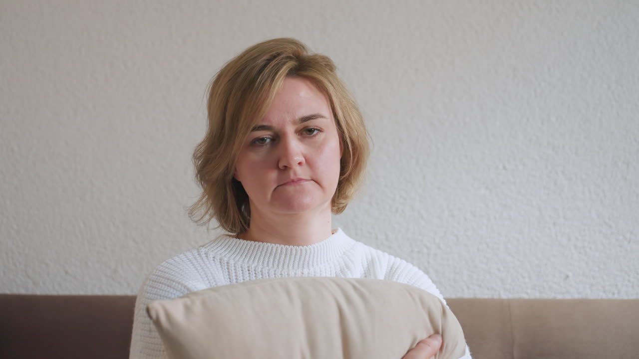 Patient seated on brown sofa holding soft beige pillow tightly, wearing white sweater, showing reflective expression in cozy therapy room with neutral walls and cushions during quiet session moment