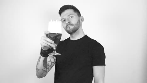 imágenes en escala de gris de un hombre tatuado levantando un vaso de cerveza con espuma en la parte superior que indica que está haciendo un brindis. tiene una cara confiada mientras mira directamente hacia adelante. el hombre tiene el cabello corto y una barba corta.