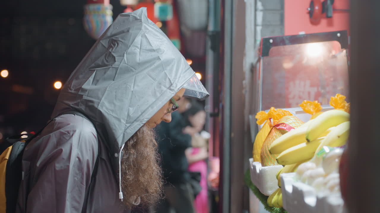 雨の夜市で果物屋台を品定めするアジア人女性。フード付きコートと眼鏡を着用し、陳列されたバナナを見ている。ランタンの光とネオンのボケ、濡れた舗道の反射が印象的で、都会の夜の群衆の中に立つ思慮深い買い物客の姿。