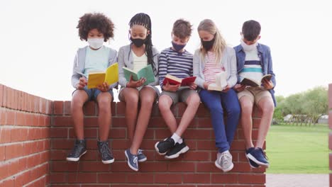 group of kids wearing face masks reading books while sitting on a brick wall