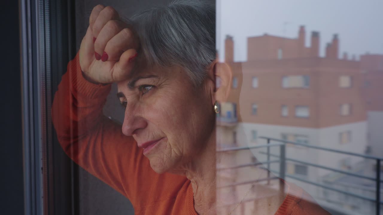 Mujer mayor pensativa mirando por la ventana con el reflejo de la ciudad.
