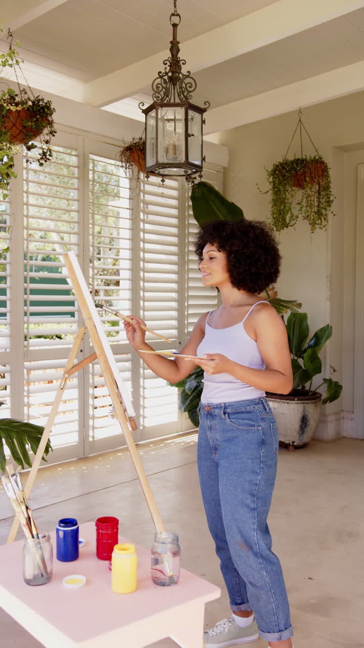 Vertical video: Painting on canvas, woman in casual outfit creating art in bright room