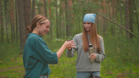 two young women in serene forest enjoy outdoor break, one pours warm drink from thermos for her friend, who collect it happily and drink from it as the smile warmly at each other