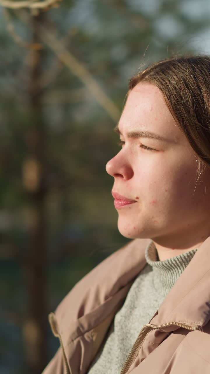 primer plano de una chica que lleva una patineta mientras camina por un parque en un día soleado. lleva una chaqueta de melocotón y una camisa interior gris, con la boca cerrada, disfrutando del ambiente tranquilo