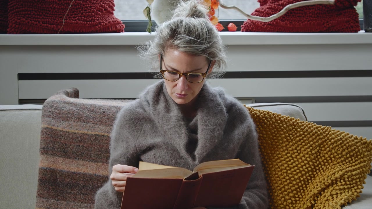 Travelling avant sur une jeune femme caucasienne à lunettes, assise sur un canapé, lisant et étudiant intensément un livre dans le salon moderne d'un appartement.