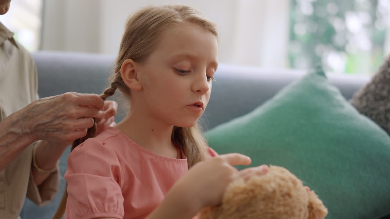 Bambina seduta sul divano. Le mani della nonna che intrecciano una treccia alla nipotina.