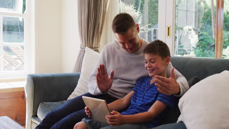 Kaukasischer-Mann-Und-Sohn-Lächeln,-Während-Sie-Einen-Videoanruf-Auf-Einem-Digitalen-Tablet-Führen-Und-Auf-Der-Couch-In-Ho-Sitzen