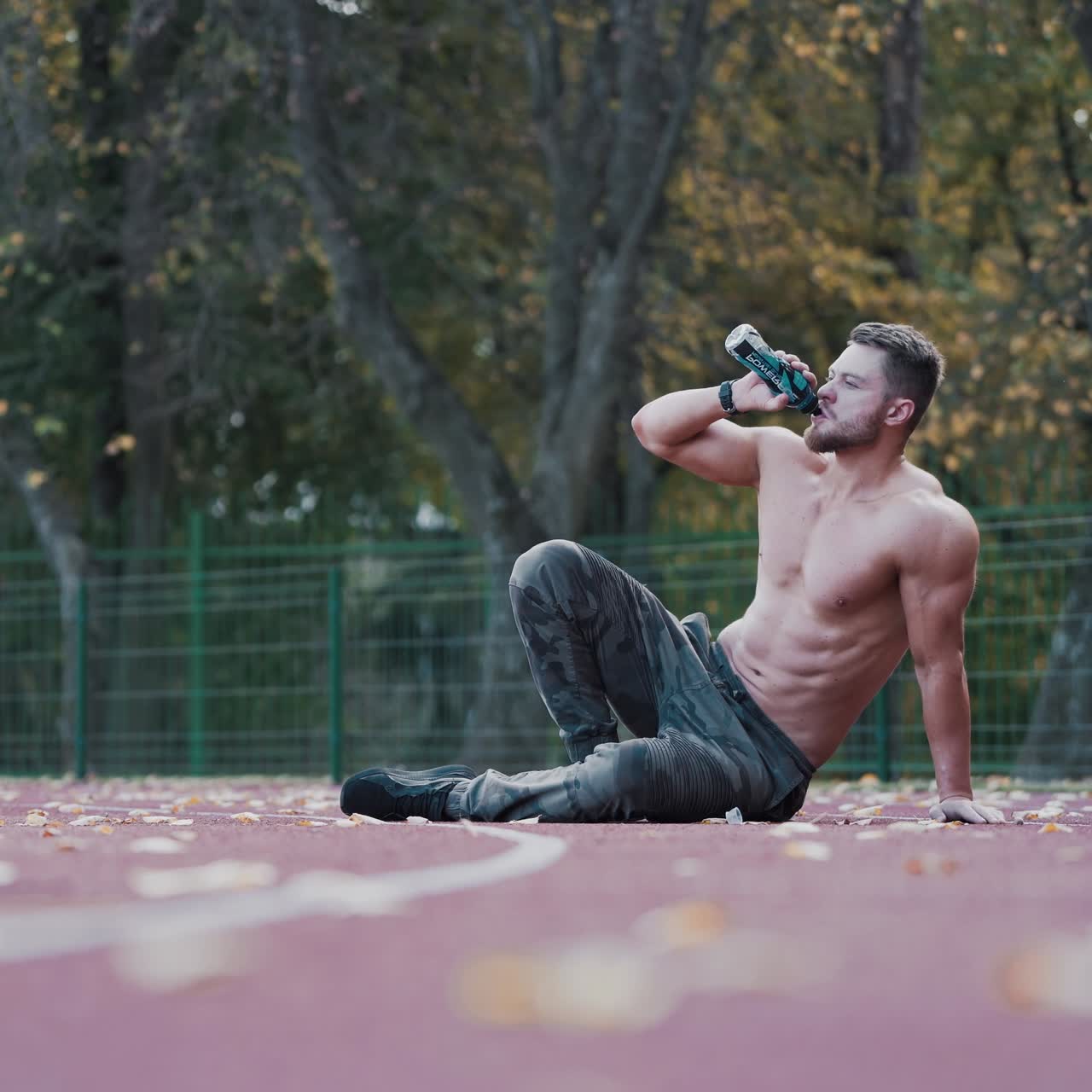 Shirtless sportsman drinks water on the stadium. Healthy athlete with muscular body sitting outdoors and having a rest after his workout. Sporty man with a bottle of water.